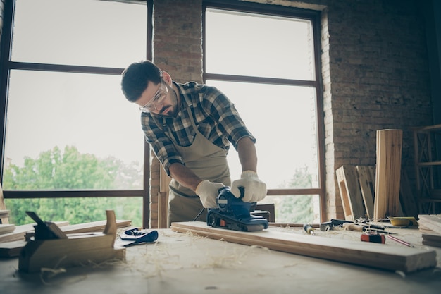 pensive man wearing checkered shirt eye glasses protecting him from sawdust working by using sander