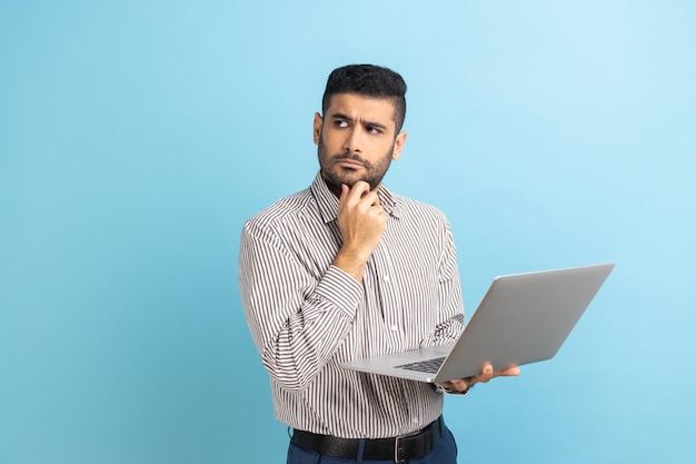 Uomo pensieroso con la barba che tiene il laptop e tiene il mento distogliendo lo sguardo pensando al progetto di business