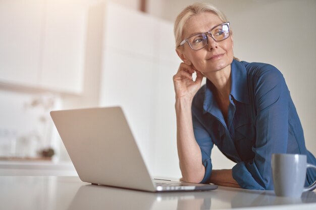 Pensive mature businesswoman looking aside working from home using laptop computer and drinking