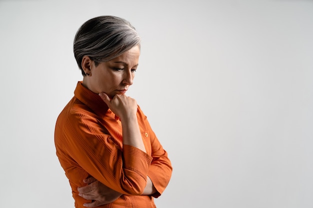 Pensive mature grey haired woman in orange shirt looks down. Pretty mid aged grey haired woman in