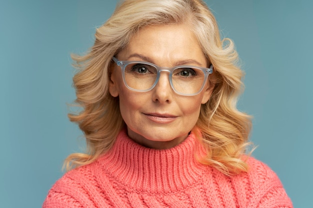 Pensive mature woman wearing stylish eyeglasses, pink  sweater isolated on blue background