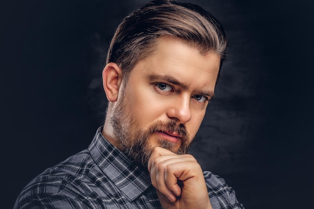 Pensive middle age man with beard and hairstyle dressed in a checkered shirt pose with hand on chin. Isolated on a dark textured background.