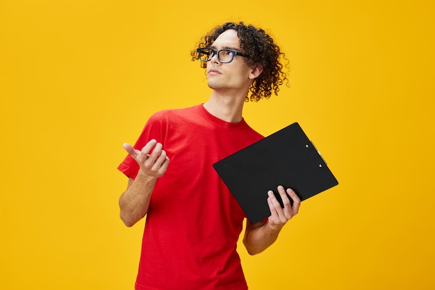 Pensive myopic young student man in red tshirt funny eyewear holds tablet folder with study notes point hand aside posing isolated on yellow background Free place for ad Education College concept