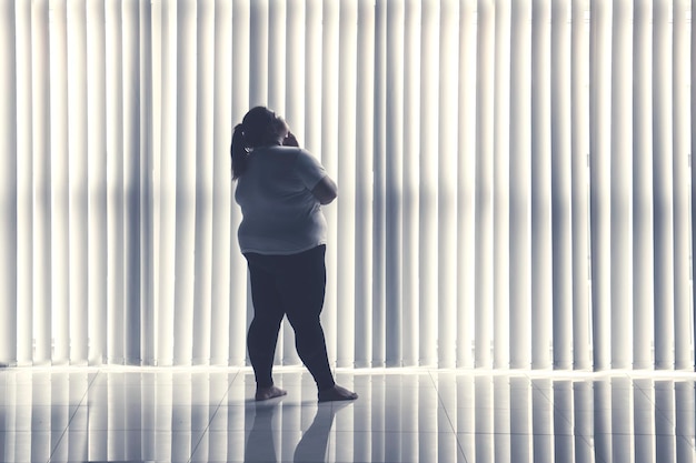 Photo pensive obese woman stands by the window