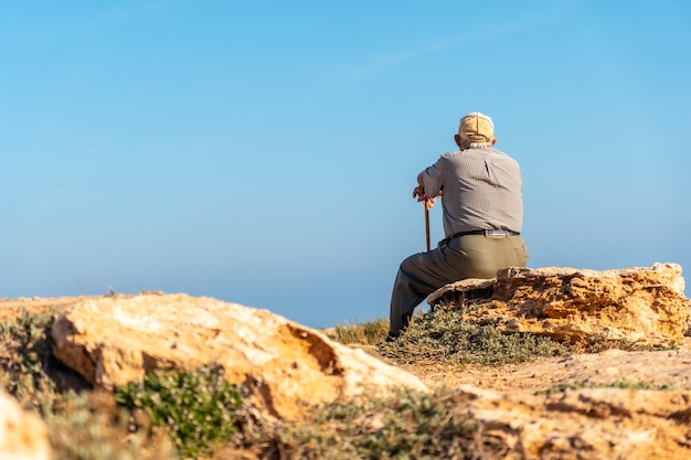 Pensive old man in beret sitting and with walking stick on the shore by the sea