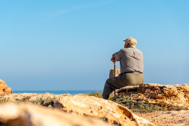 Pensive old man in beret sitting and with walking stick on the shore by the sea