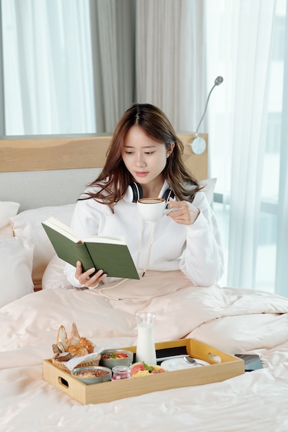 Pensive pretty young woman drinking cup of cappuccino and reading reading interesting book when sitting on bed at home