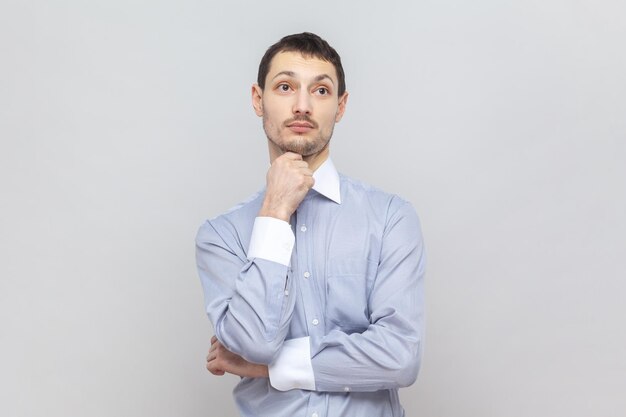 Pensive puzzled young adult man standing holding chin looking away with thoughtful expression