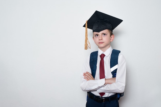 Pensive schoolboy wearing students hat and crossed his arms. Children education concept. White wall, copy space