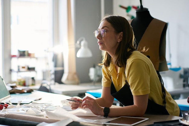 Pensive seamstress in eyeglasses sitting by table with sewing patterns