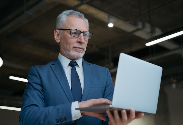 Pensive senior businessman using laptop, typing, searching online, reading news in office