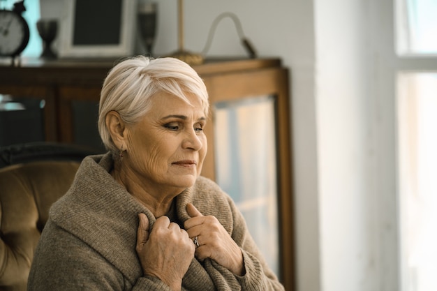 Donna senior pensierosa che si avvolge in un maglione caldo che siiting sul sofà accogliente