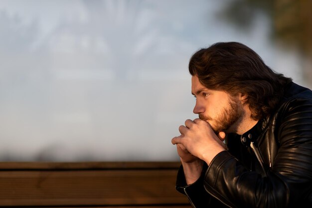 Задумчивый серьезный молодой бородатый мужчина носит черную кожаную куртку сидит и думает на открытом воздухе