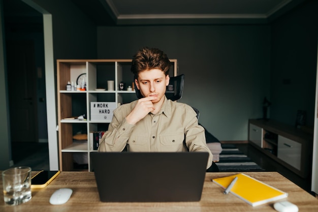 Задумчивый парень без рубашки, работающий в спальне за столом на ноутбуке, смотрит на экран и думает с серьезным лицом Молодой человек работает удаленно из дома на ноутбуке