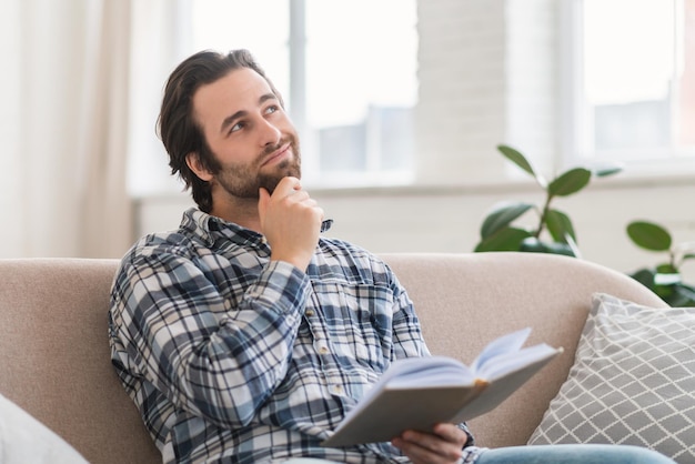 Pensive smart millennial european guy in shirt with stubble with book or diary reads or dreaming on sofa