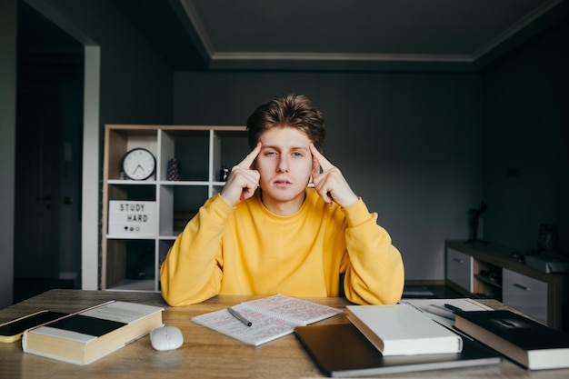 Pensive student sits at a desk at home with books and a notebook looks focused at the camera