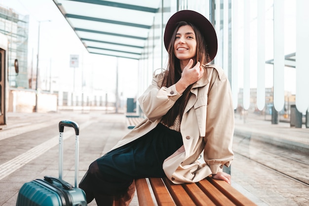 Фото Задумчивая стильная девушка в пальто и шляпе сидит на остановке в ожидании общественного транспорта