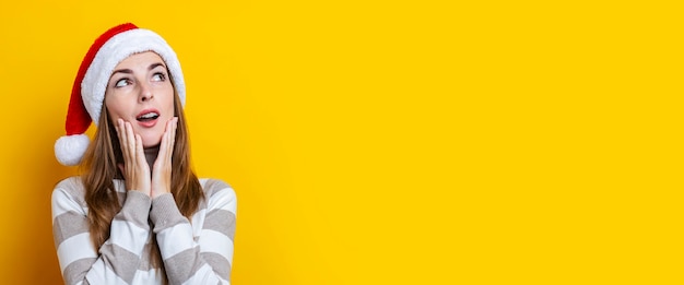 Pensive surprised young woman in Santa Claus hat on a yellow background. Banner.