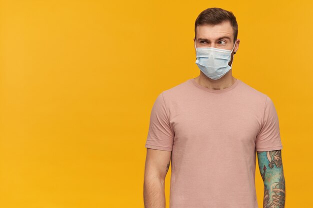 Pensive suspicious young bearded tattooed man in virus protective mask on face against coronavirus with raised brow standing and looking away over yellow wall