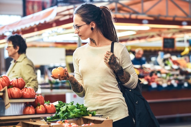 Pensive tattoed girl is carefully choosing tomatoes on a local farmer's market.