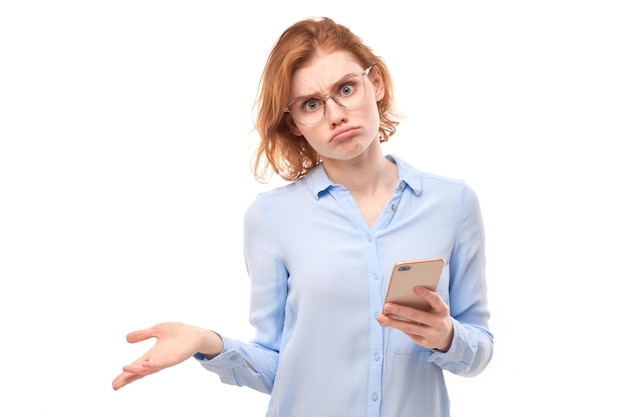 Рыжая девушка с задумчивым мышлением, держащая смартфон с расстроенным лицом в очках и деловой рубашке, изолирована на белом студийном фоне