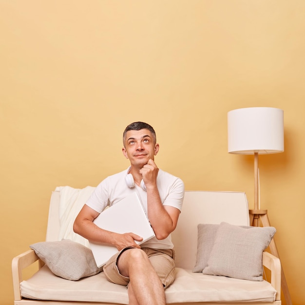 Pensive thoughtful adult man wearing white tshirt and shorts sitting on sofa with closed laptop against beige wall looking upwards at advertisement area copy space for promotion