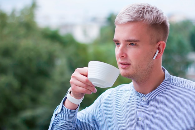 Pensive thoughtful guy drinking drink, tea or coffee from a cup. Young man with gadgets in a shirt listening to music in wireless earphones outdoor.