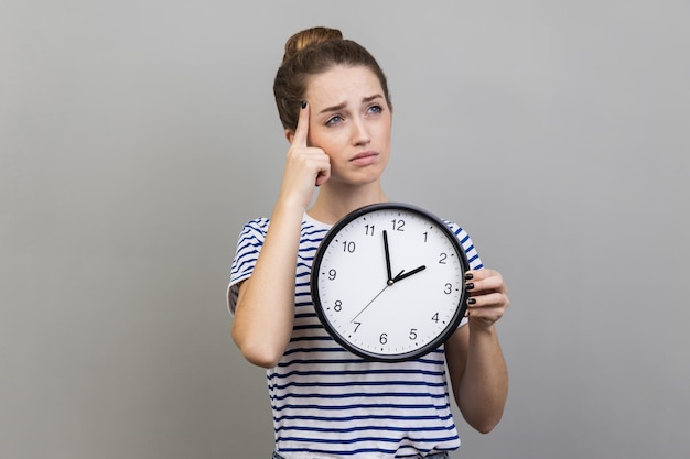 Pensive thoughtful woman wearing striped tshirt holding wall clock thinking about deadline