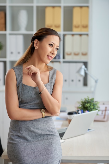 Pensive Vietnamese female executive in gray dress leaning on table with office supplies and looking away