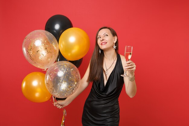 Pensive woman in black dress celebrate make wish looking up hold glass of champagne air balloons isolated on red background. St. Valentine's Day, Happy New Year, birthday mockup holiday party concept.