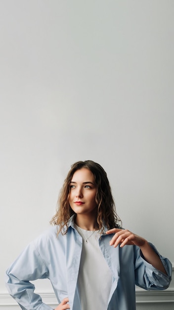 A pensive woman in blue shirt reminiscing about past love smiling yet with a touch of sadness standing against a white wall and playing with hair