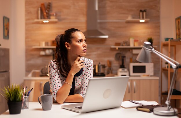 밤늦게 노트북 작업을 하는 동안 집에 있는 부엌에서 생각에 잠긴 여성. 자정에 현대 기술을 사용하는 직원은 업무, 비즈니스, 바쁜, 경력, 네트워크, 생활 방식, 무선을 위해 초과 근무를 합니다.
