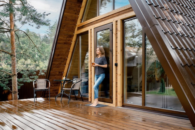 Una donna pensosa si trova sulla terrazza di un bungalow di legno con un bicchiere di tisana