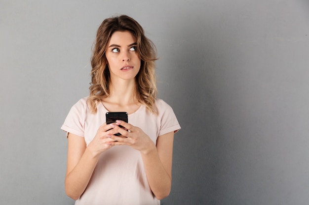 Pensive woman in t-shirt holding smartphone and looking away over grey