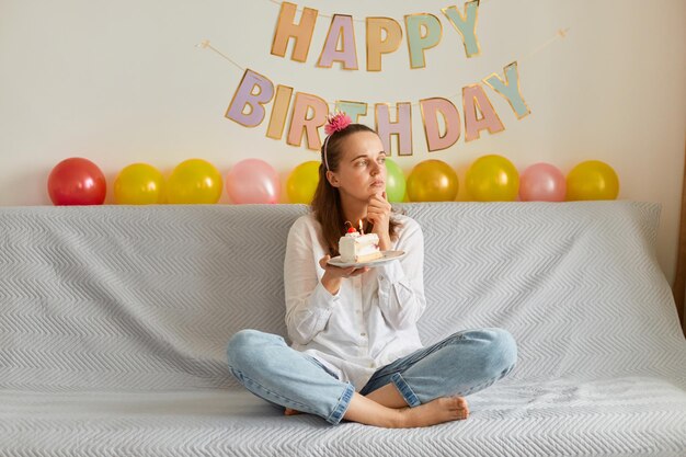 Donna pensierosa che indossa una camicia bianca e jeans seduta sul divano con le gambe incrociate, tenendo la torta con la candela, guardando da parte e esprimendo un desiderio, festeggiando il compleanno, decorazione festiva sullo sfondo.