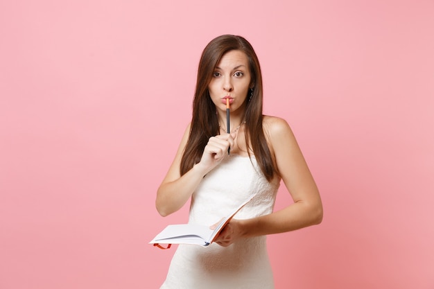 Pensive woman in white dress holding pencil near lips search ideas, writing notes in notebook