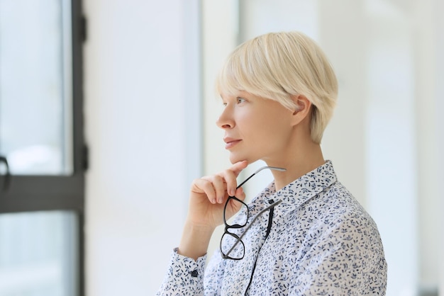 Pensive woman with short blonde hair looks out window holding glasses under chin elegant