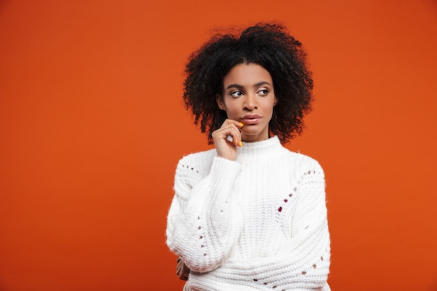 Pensive young african woman wearing sweater standing isolated over red wall, looking away