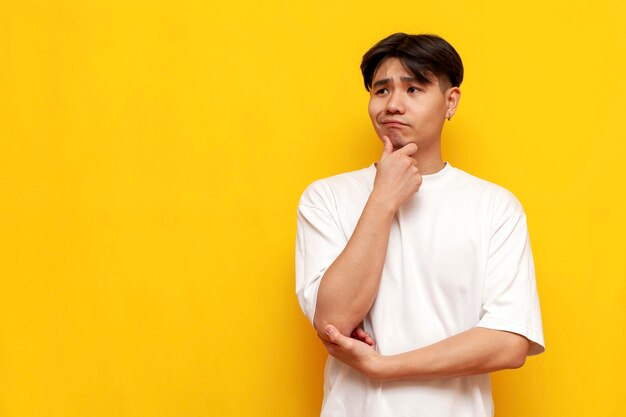 pensive young asian guy in white tshirt thinks and plans on yellow isolated background