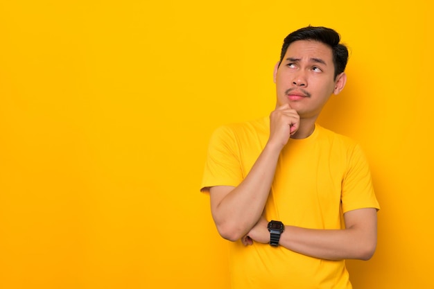 Pensive young Asian man in casual tshirt looking up at copy space and touching chin thinking about interesting offer isolated on yellow background People lifestyle concept