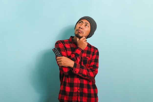 Pensive young Asian man choosing movie to watch while holding TV remote isolated on blue background