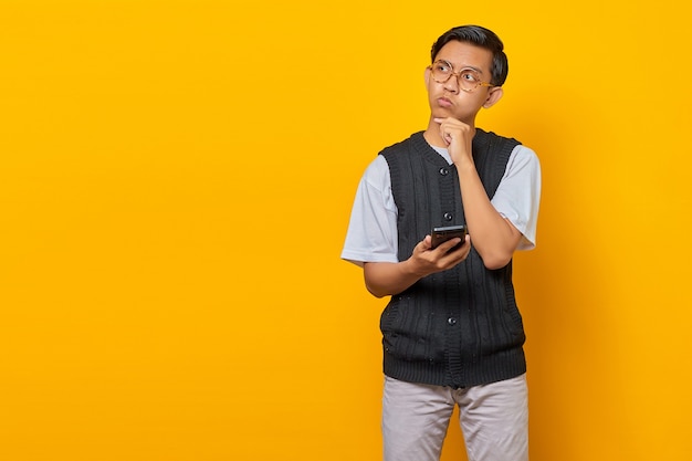 Pensive young Asian man using mobile phone with fingers on chin and looking aside