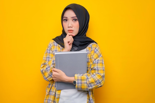 Pensive young asian muslim student holding book looks seriously\
thinking about a question isolated on yellow background back to\
school in high school college concept