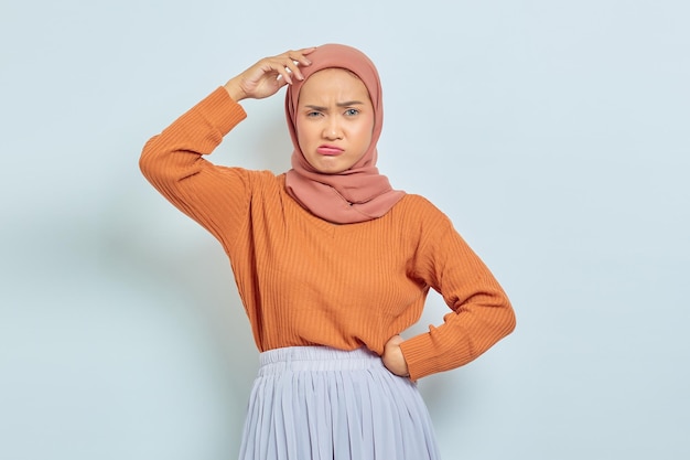 Задумчивая молодая азиатская мусульманка в коричневом свитере и хиджабе выглядит серьезно, размышляя над вопросом, изолированным на белом фоне