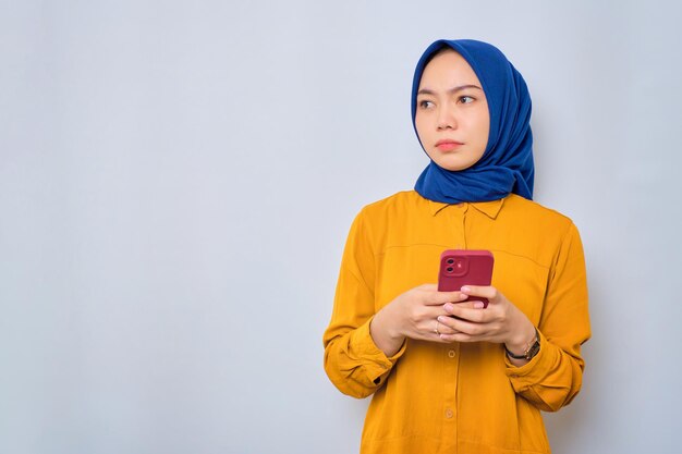 Pensive young asian muslim woman dressed in orange shirt\
holding mobile phone and looking aside at copy space thinking about\
interesting offer isolated over white background