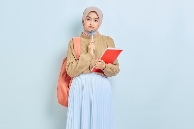 Задумчивая молодая азиатская студентка-мусульманка в коричневом свитере и рюкзаке держит книгу и ручку, думая о чем-то изолированном на белом фоне обратно в школу