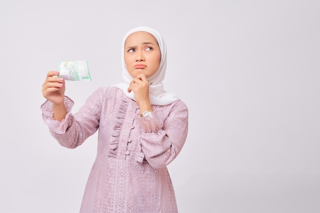 Pensive young Asian Muslim woman wearing hijab and purple dress holding fan of cash money and touching chin isolated on white studio background