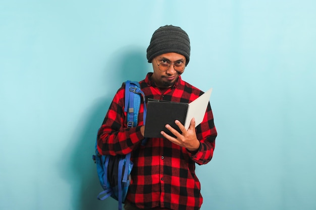 Pensive young Asian student man is thinking while holding book and standing against blue background