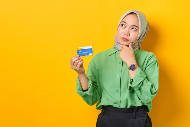 Pensive young asian woman in a green shirt holding credit card and looking away on yellow background