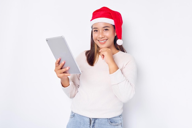 Pensive young Asian woman in a hat Christmas using a digital tablet and touching her cheek isolated over white background Indonesian people celebrate Christmas concept